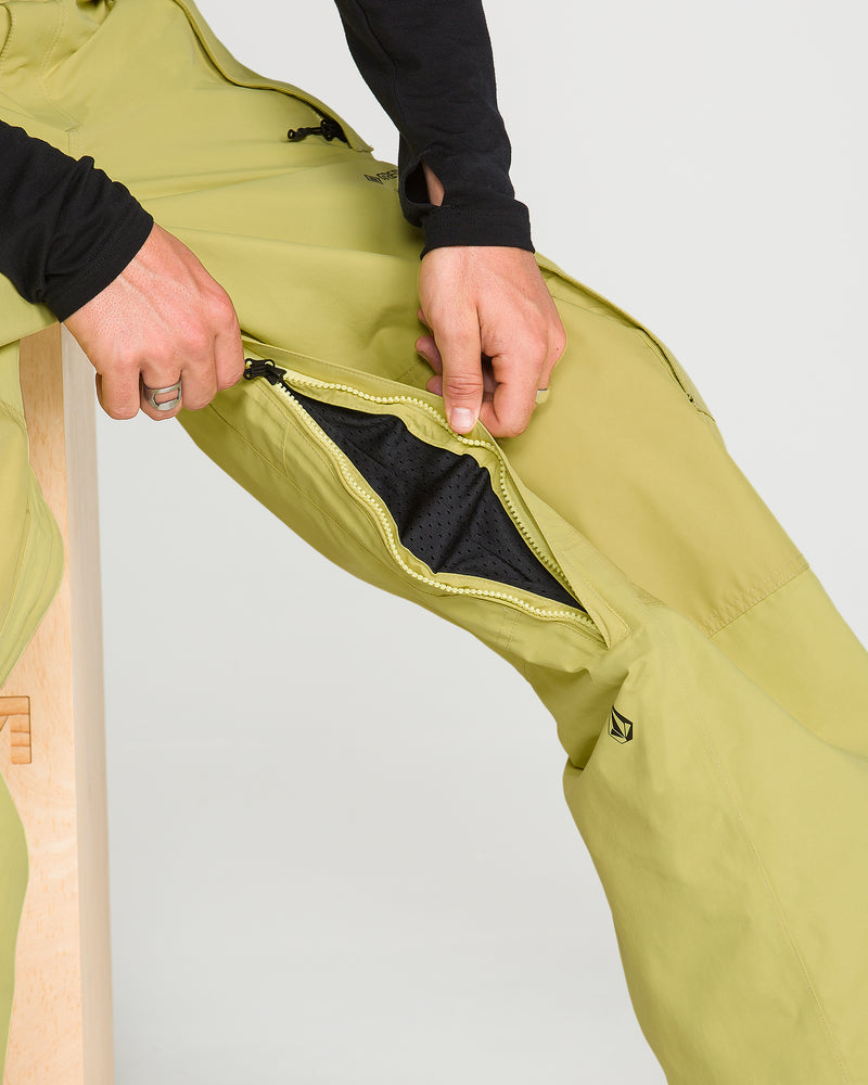 Wearing Volcom Men’s Longo Gore-Tex Pants in a light green shade, a person unzips the side leg vent to reveal black mesh fabric beneath—a signature detail inspired by Arthur Longo’s style. As they sit comfortably on a wooden block, these pants offer breathable protection in any condition.