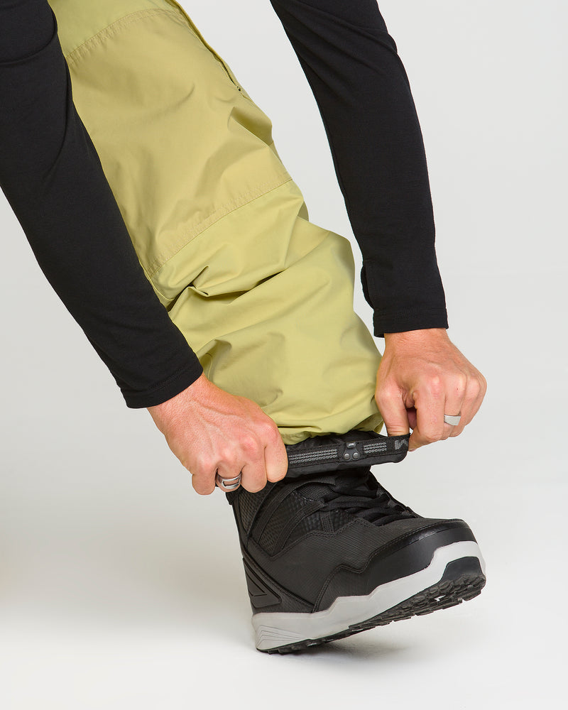 Wearing Volcom Men’s Longo Gore-Tex Pants in yellow-green and a black long-sleeve shirt, a person pulls the cuff of their pants over a black snow boot, preparing for outdoor activity.
