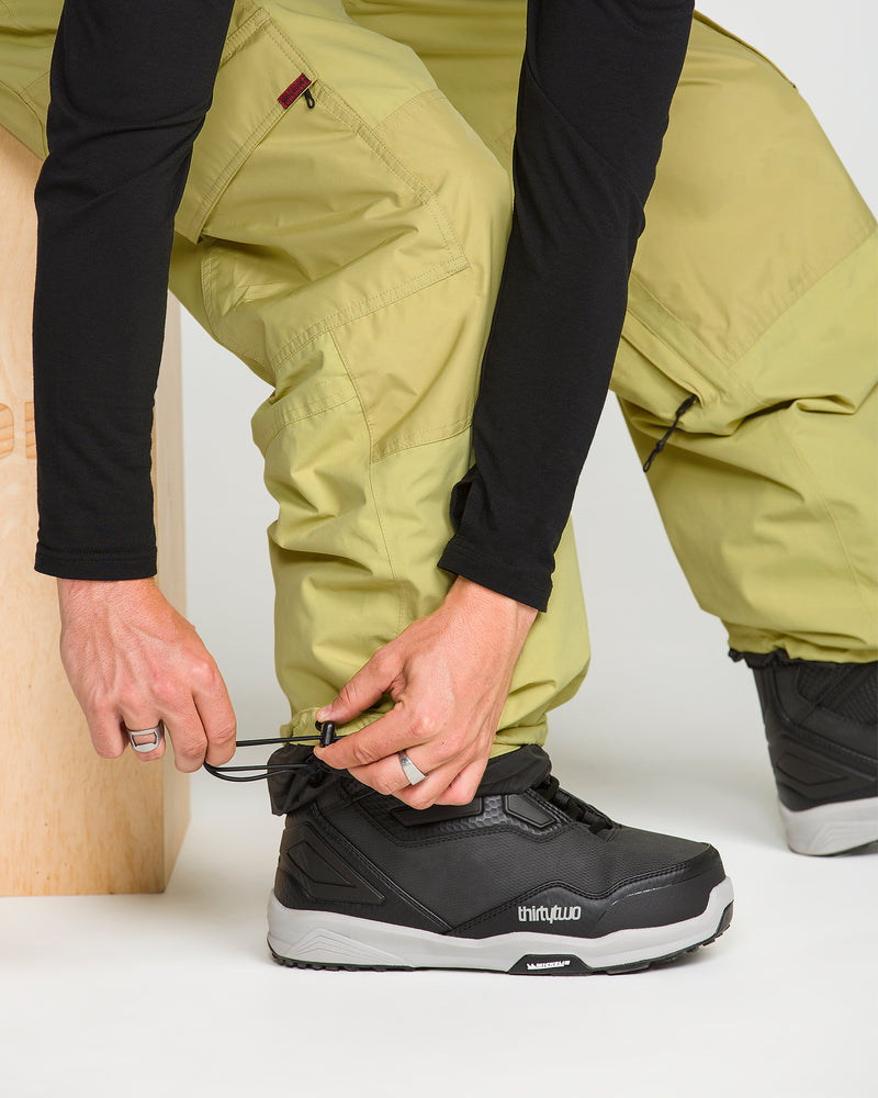 A person wearing yellow-green Volcom Men’s Longo Gore-Tex Pants and a black long-sleeve shirt ties the laces of black ThirtyTwo snowboarding boots.