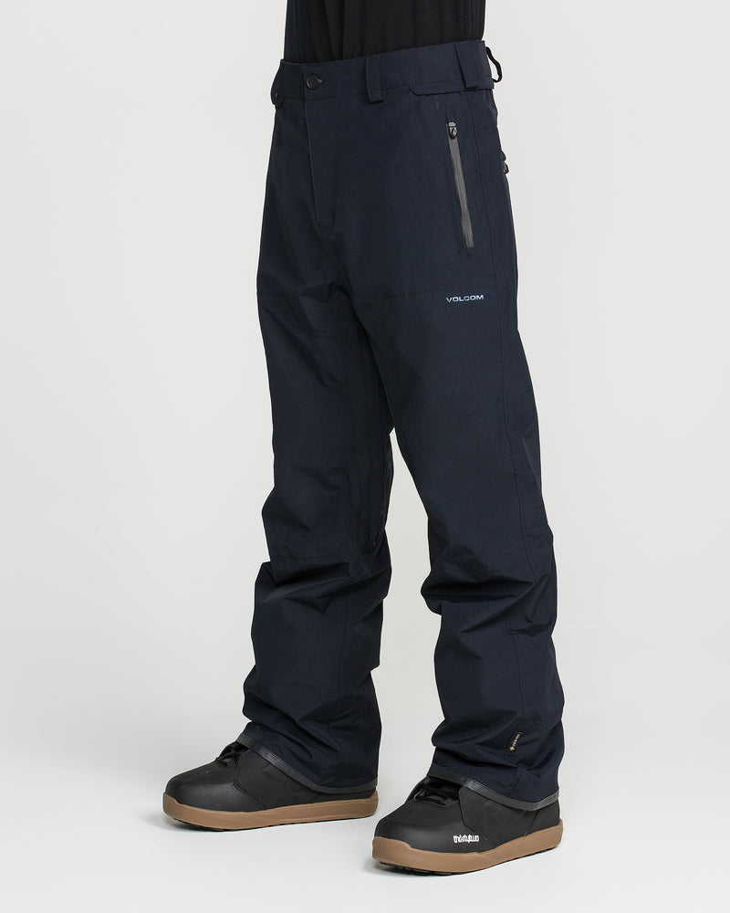 A person is shown from the waist down, standing on a white background, wearing black Volcom Men’s L Gore-Tex Pants and black winter boots. The pants are made from recycled, PFAS-free GORE-TEX fabric and feature a zippered pocket with a subtle Volcom logo on the thigh.