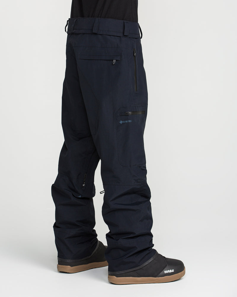 A person is wearing loose-fitting, dark blue Volcom Men’s L Gore-Tex Pants with black winter boots. These pants feature zippered pockets and are made from waterproof, PFAS-free GORE-TEX material, making them suitable for cold or snowy weather.