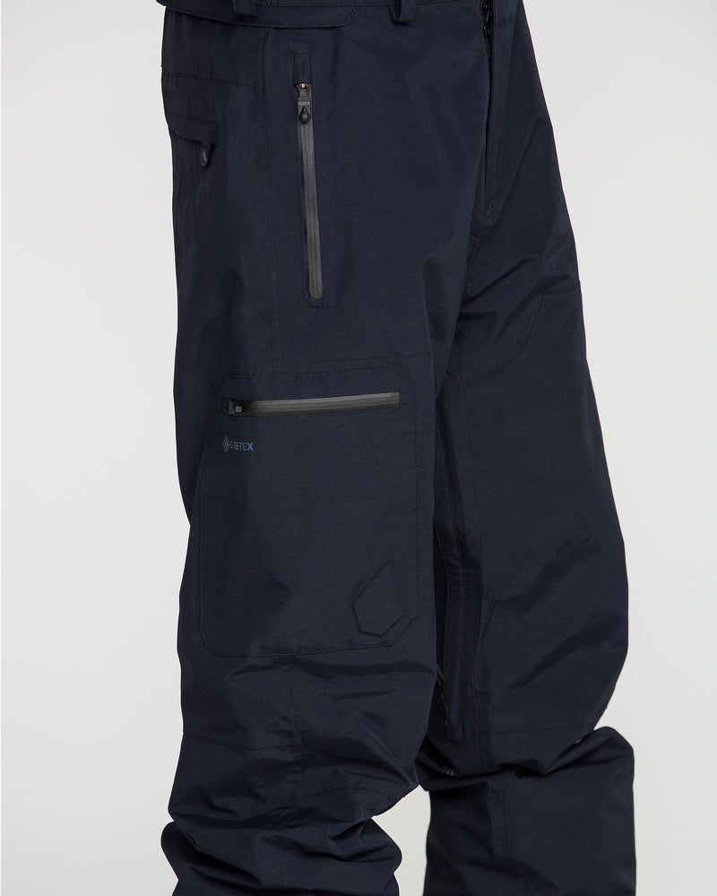 Close-up of a person wearing Volcom Men’s L Gore-Tex Pants in dark navy, featuring zippered pockets and a visible recycled GORE-TEX logo, with the upper legs shown against a plain background.
