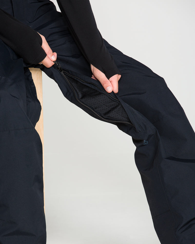 A person unzips the mesh-lined vent on the side of a pair of black Volcom Men’s L Gore-Tex Pants while sitting on a wooden stool.