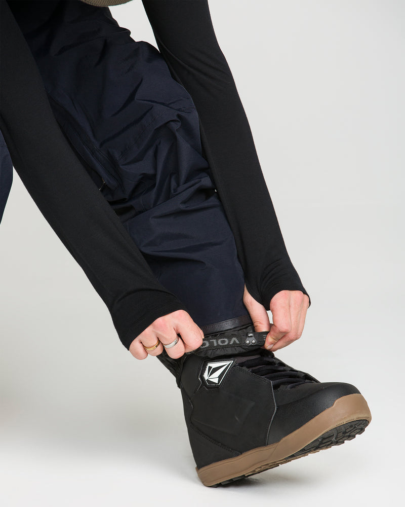 Wearing Volcom Men’s L Gore-Tex Pants and a long-sleeve black shirt, a person pulls the cuff over the top of a black snowboarding boot with a brown sole.