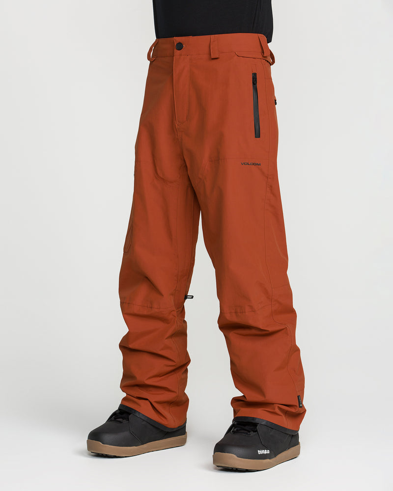A person is wearing Volcom Men’s L Gore-Tex Pants in a loose fit and rust color, featuring a zippered pocket on the right side. The pants are made from recycled, PFAS-free GORE-TEX material. The outfit is completed with black boots and a black shirt against a plain white background.