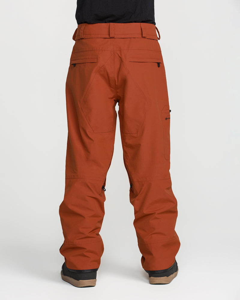 A person wearing rust-colored Volcom Men’s L Gore-Tex Pants, which are PFAS-free, and black boots is standing facing away from the camera against a plain white background.