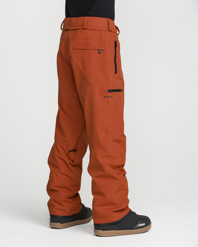 A person is shown from the side and back wearing burnt orange Volcom Men’s L Gore-Tex Pants—crafted from recycled, PFAS-free GORE-TEX fabric—and black boots. The pants feature zippered pockets, belt loops, and visible seams against a plain white background.