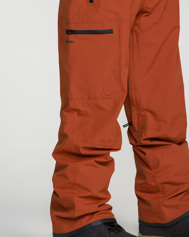 Close-up of a person wearing Volcom Men’s L Gore-Tex Pants in a rust color, featuring zippered pockets and paired with black boots. The durable fabric is crafted from recycled GORE-TEX, making these pants both ideal for hiking and outdoor activities and an environmentally conscious choice.
