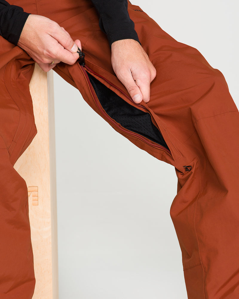 A person unzips a vent along the thigh of rust-colored Volcom Men’s L Gore-Tex Pants, revealing a black mesh lining beneath.