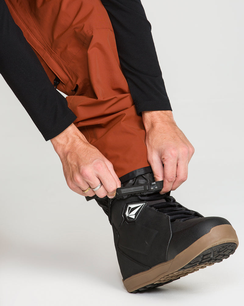 A person wearing Volcom Men’s L Gore-Tex Pants in rust is tightening the laces of a black snowboard boot with a brown sole. Only their hands and lower legs are visible against a plain background.