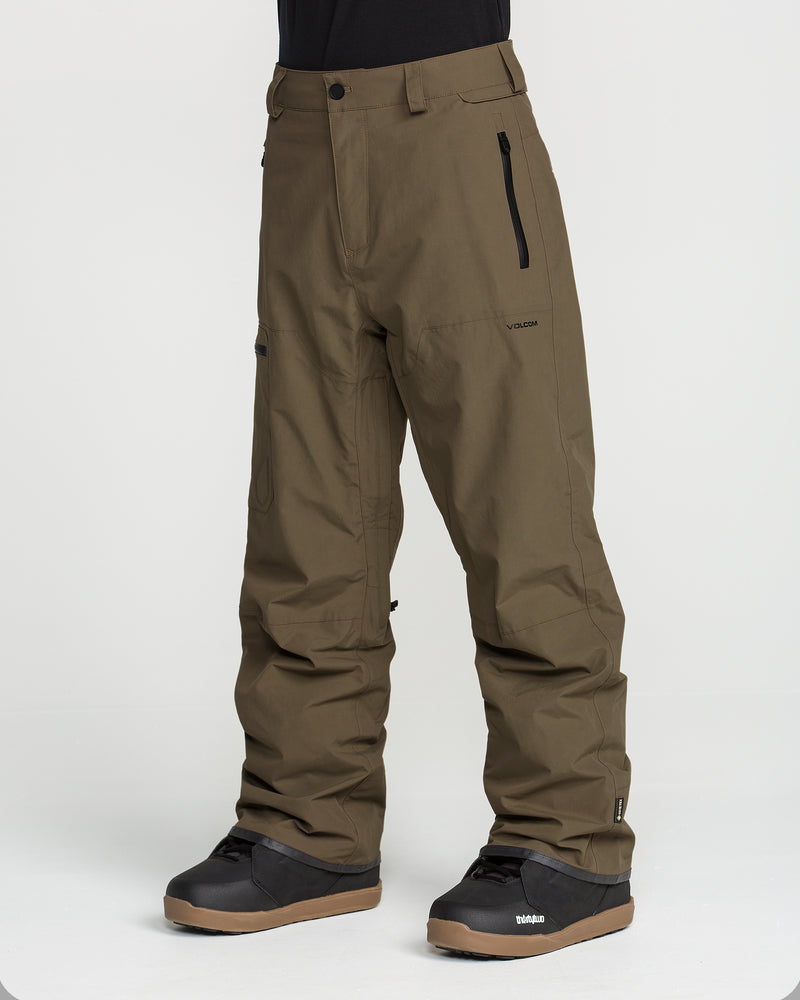 A person is shown from the waist down, standing against a plain white background, wearing Volcom Men’s L Gore-Tex Pants in olive green. These recycled, PFAS-free snow pants feature zippered pockets and a relaxed fit, paired with black boots.