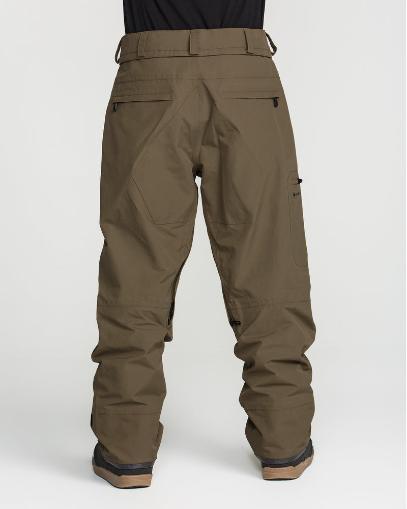 A person is shown from behind against a plain white background, wearing Volcom Men’s L Gore-Tex Pants in olive green, crafted from recycled, PFAS-free GORE-TEX fabric for sustainable style. The loose-fitting cargo pants are paired with brown shoes.
