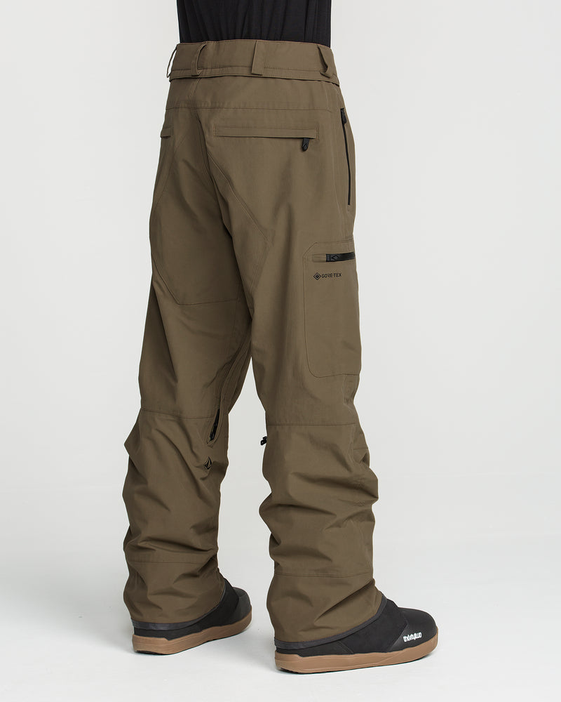 A person stands facing away, wearing Volcom Men’s L Gore-Tex Pants in olive green, featuring multiple pockets and zippers, paired with black and brown snow boots. The background is plain white.