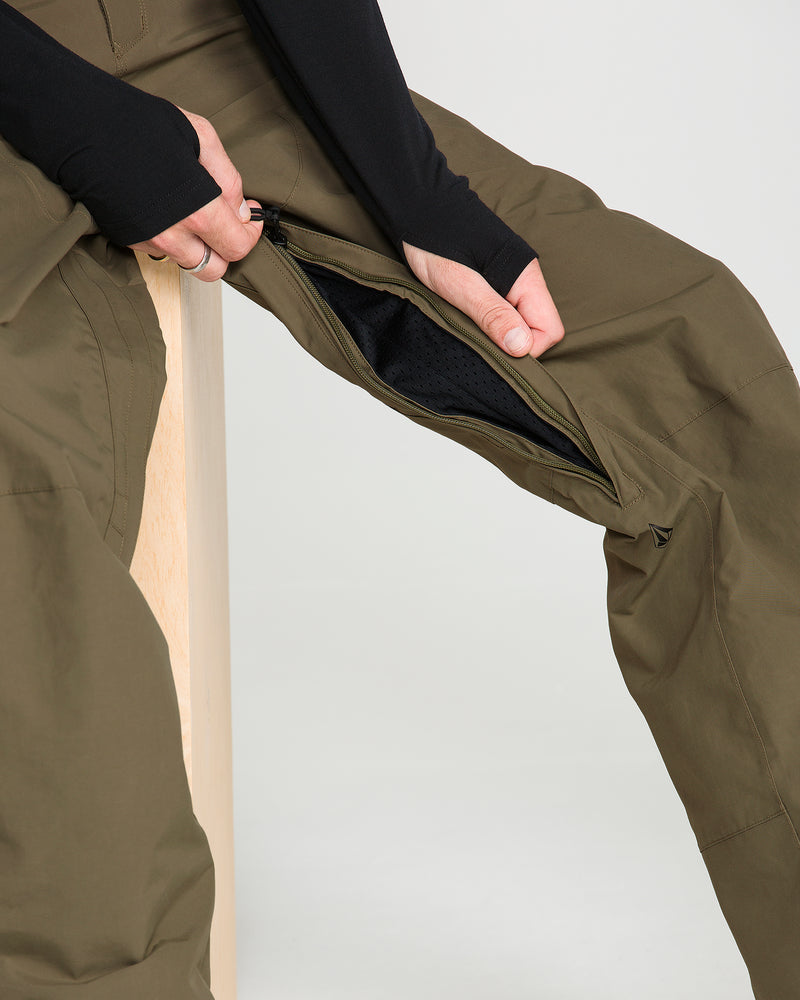 A person sits and unzips a vent on the thigh of the Volcom Men’s L Gore-Tex Pants in olive green, revealing a black mesh lining inside while holding the zipper.