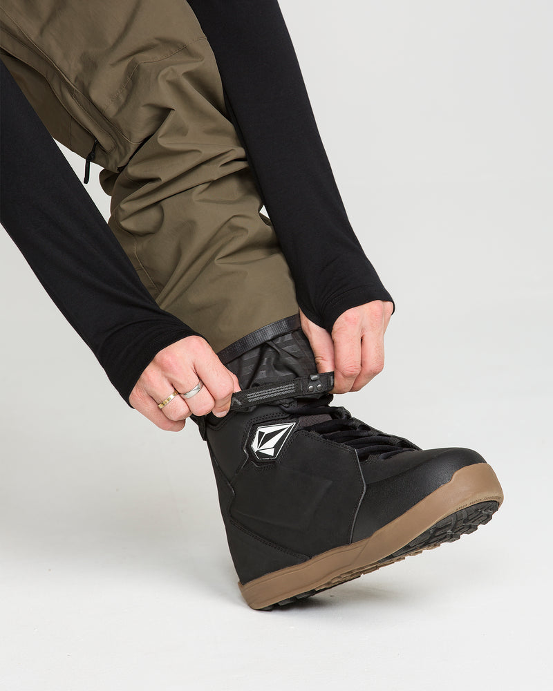 A person wearing Volcom Men’s L Gore-Tex Pants in brown adjusts the strap on a black snowboarding boot with a brown sole. Only the lower legs and hands are visible.
