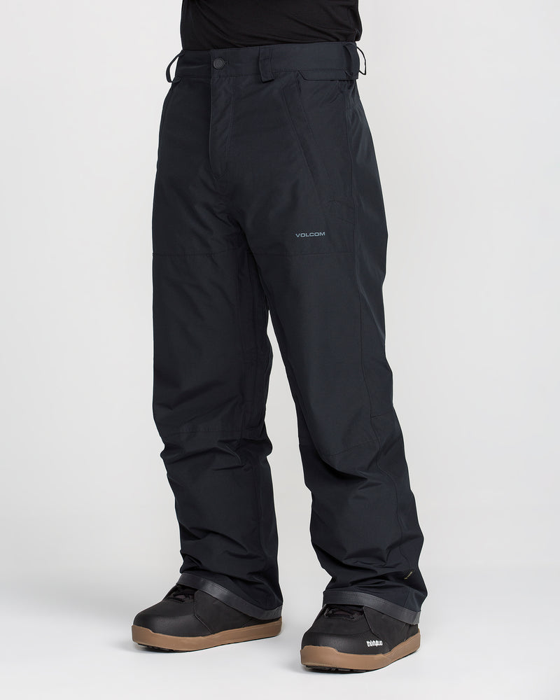 A person wearing black Volcom Men’s Dua Gore-Tex Pants with a breathable lining, black snowboard boots, and a black shirt stands against a plain white background.