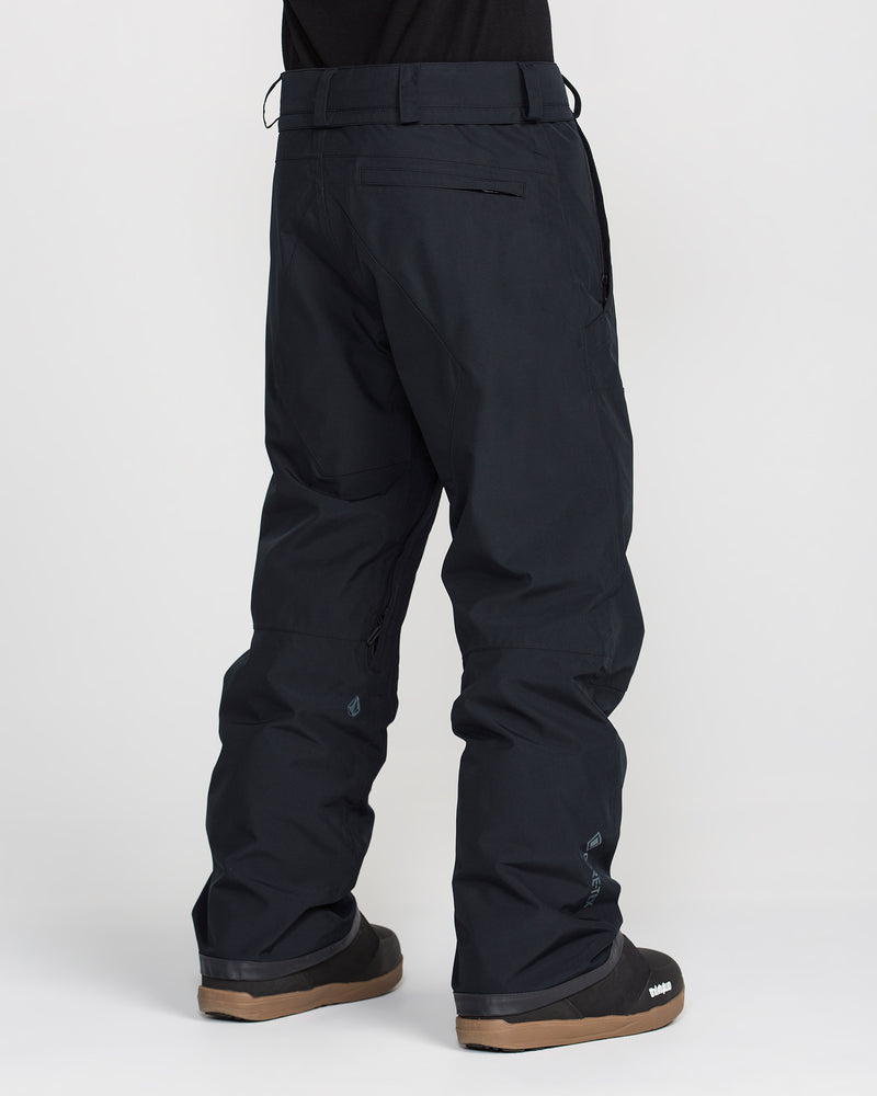 A person is wearing Volcom Men’s Dua Gore-Tex Pants in a relaxed fit paired with black boots featuring brown soles, standing against a plain white background. The rear view emphasizes the pants’ detailed pockets and seams, while showcasing the breathable lining that enhances comfort in these waterproof pants.