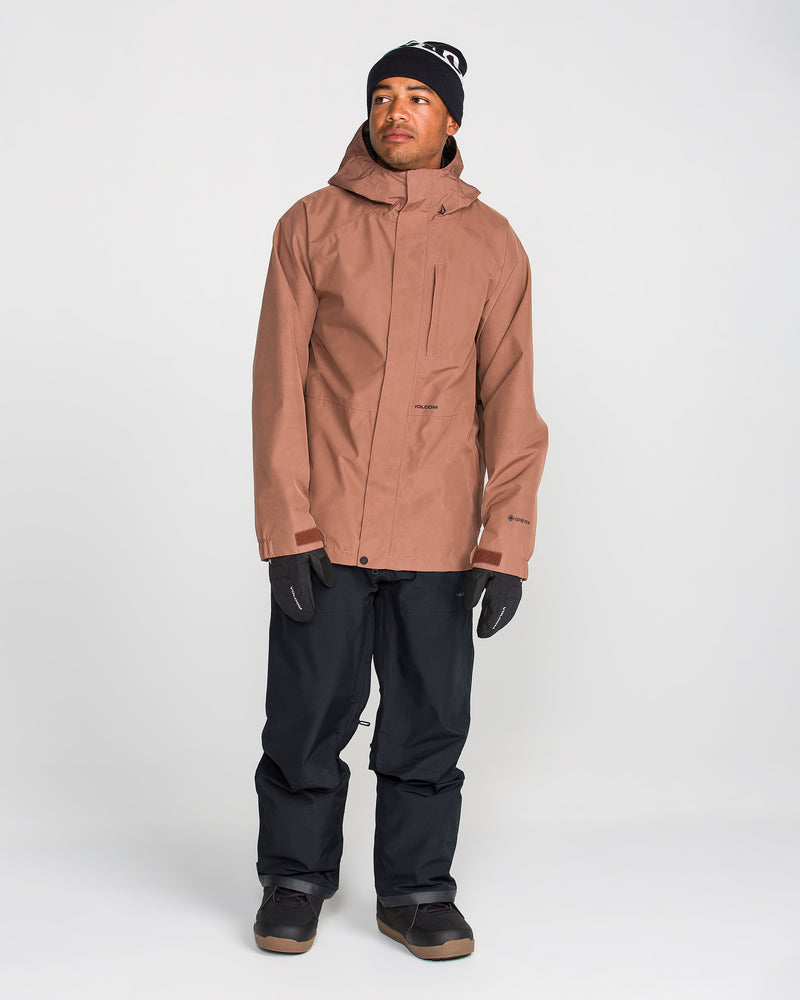 A person wearing a brown hooded jacket, Volcom Men’s Dua GORE-TEX Pants with breathable lining in black, black gloves, and a black beanie stands facing slightly left against a plain white background.