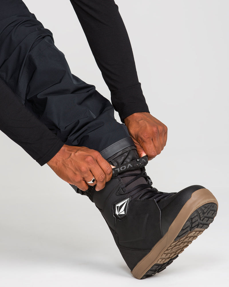 Wearing Volcom Men’s Dua Gore-Tex Pants, a person adjusts the cuff over a black snow boot with tan sole while preparing for outdoor activity.