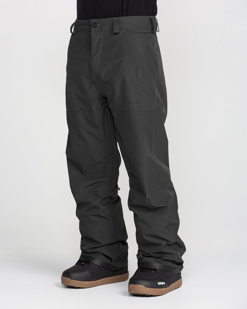 A person wearing black Volcom Men’s Dua GORE-TEX Pants and black snow boots with tan soles stands against a plain white background. The PFAS-free pants feature zippered pockets, a breathable lining, and reinforced knee areas.