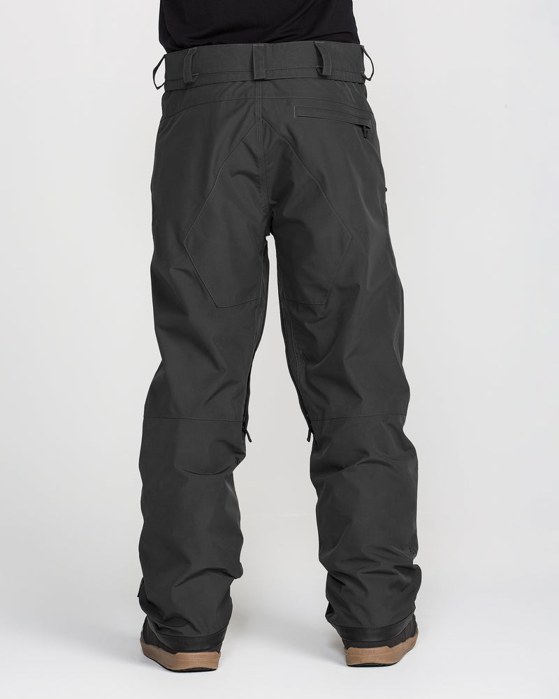 A person is shown from behind, wearing Volcom Men’s Dua Gore-Tex Pants in loose-fitting dark gray, featuring visible seams, pockets, and belt loops. The pants have a breathable lining and are PFAS-free, styled casually for outdoor use. The look is completed with brown shoes against a plain light background.