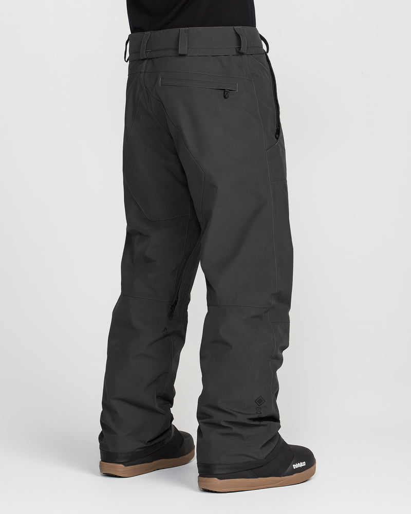 A person is wearing the Volcom Men’s Dua GORE-TEX Pants in black along with black winter boots, standing against a white background. The snow pants feature side and back zippered pockets and are designed with a breathable lining for added comfort. The image highlights the lower body.