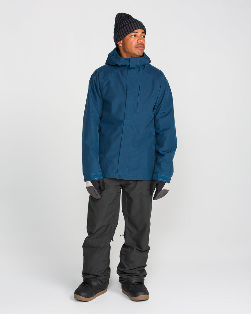 A person wearing a dark blue jacket with a breathable lining, black beanie, gray gloves, Volcom Men’s Dua GORE-TEX Pants in black, and black boots stands looking to the side against a plain white background.