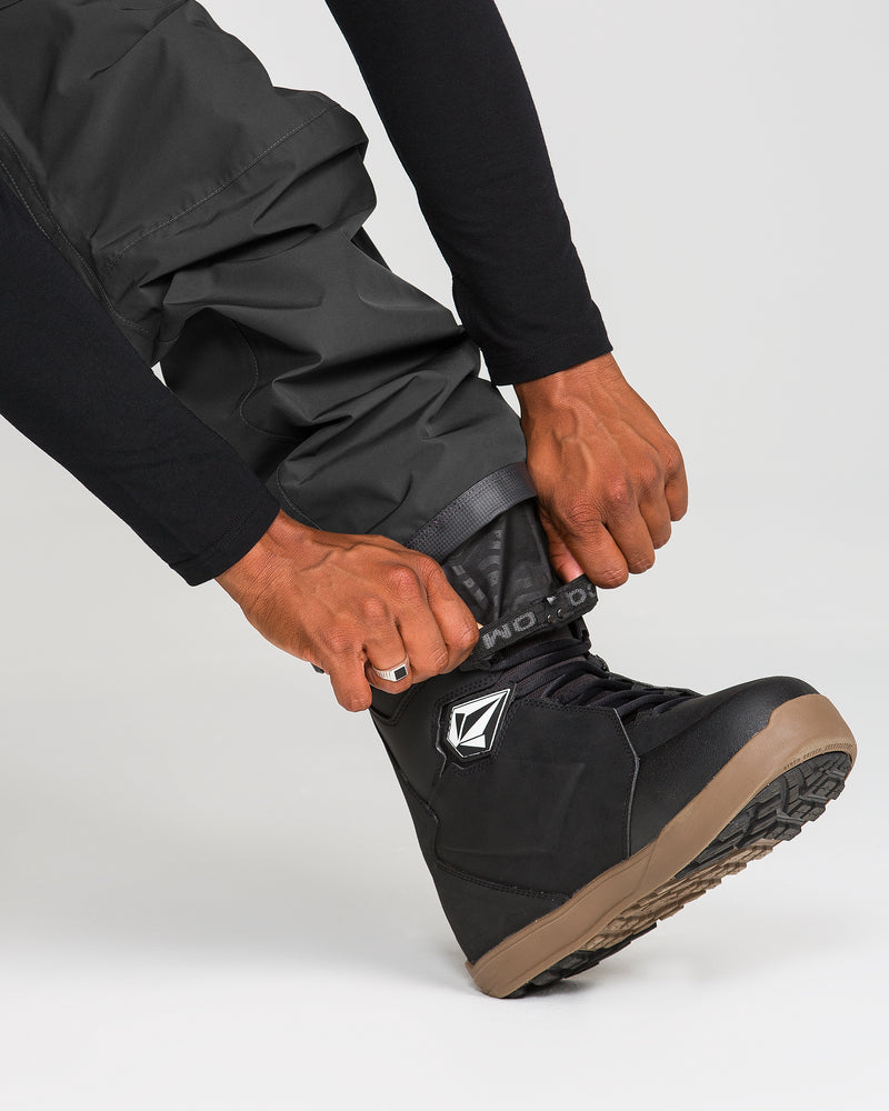 A person wearing Volcom Men’s Dua Gore-Tex Pants in black fastens the strap on a black snowboarding boot with a brown sole.
