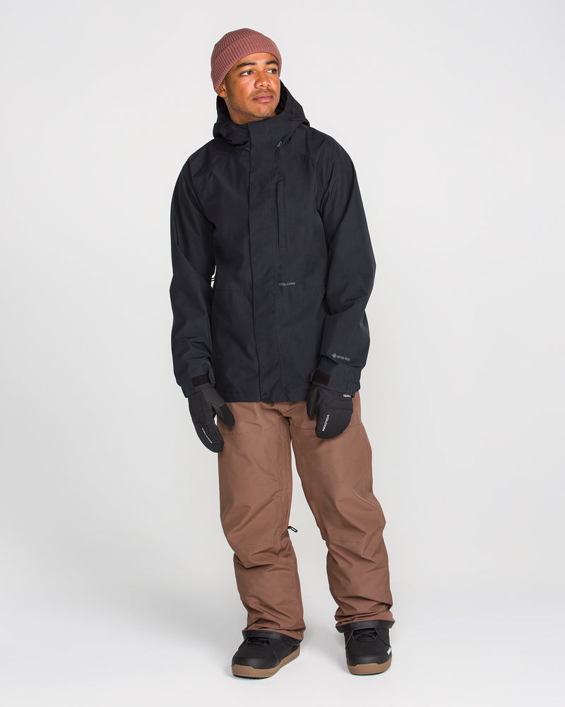 A person stands against a plain white background wearing a black hooded jacket, Volcom Men’s Dua GORE-TEX Pants with breathable lining, black gloves, black shoes, and a maroon knit beanie.