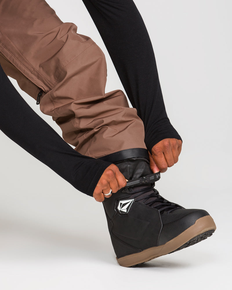 A person wearing Volcom Men’s Dua Gore-Tex Pants in brown and a black long-sleeve shirt fastens the strap on a black snowboard boot with a white logo, against a plain background.