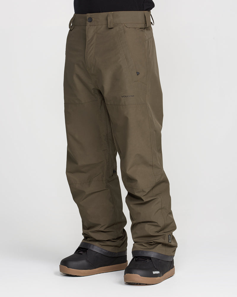 A person is wearing Volcom Men’s Dua Gore-Tex Pants in olive green, paired with black snow boots, standing against a plain white background. The pants offer waterproof protection with GORE-TEX technology, a button closure, breathable lining, and convenient side pockets.