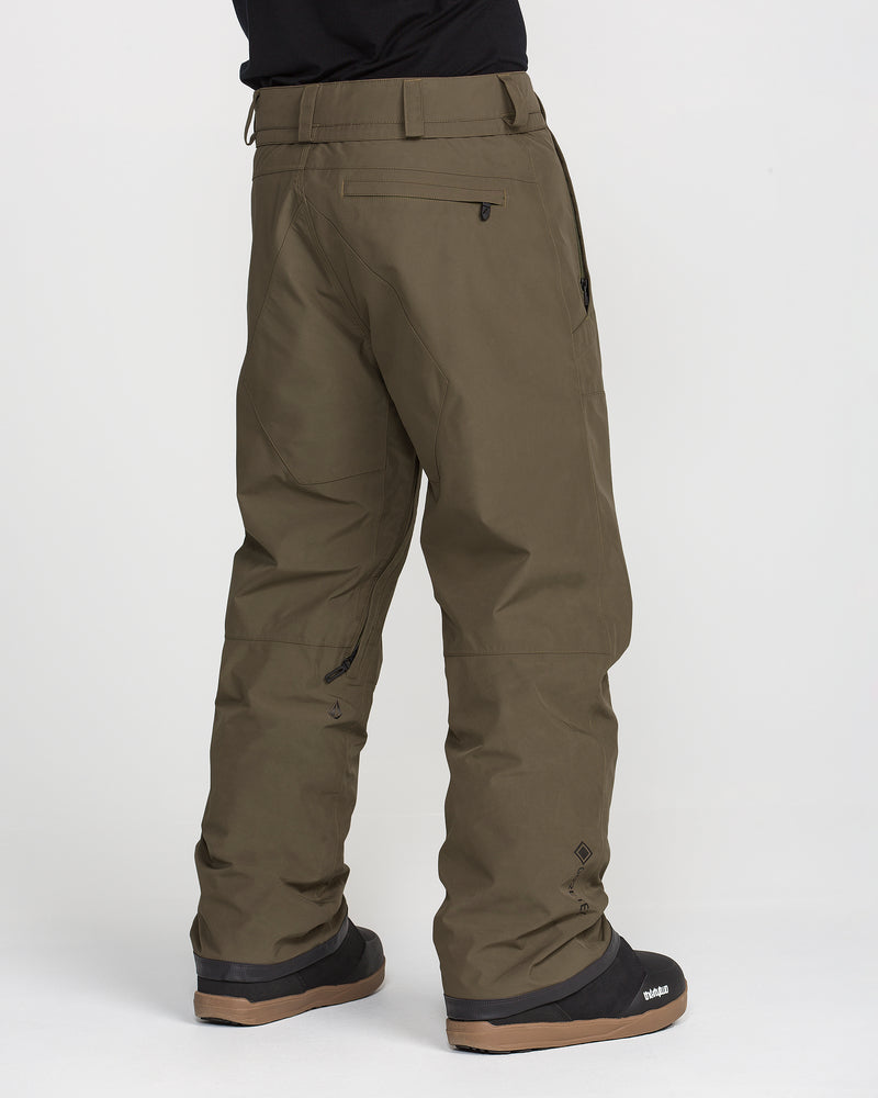 A person is shown from the back at a slight angle, standing on a white background, wearing Volcom Men’s Dua GORE-TEX Pants in olive green along with black snow boots. The pants offer waterproof protection, a zippered pocket, breathable lining, and reinforced fabric at the cuffs.