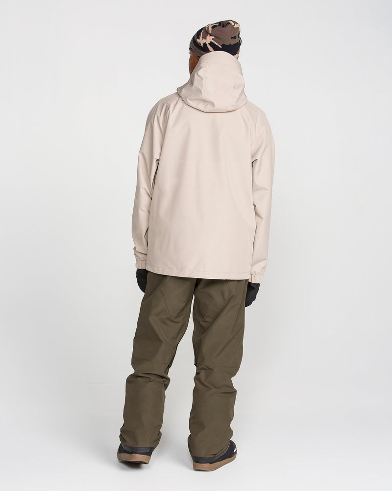 A person stands facing away, wearing a beige hooded jacket with waterproof protection, Volcom Men’s Dua GORE-TEX Pants in olive green featuring a breathable lining, black gloves, brown boots, and a camouflage beanie, against a plain white background.