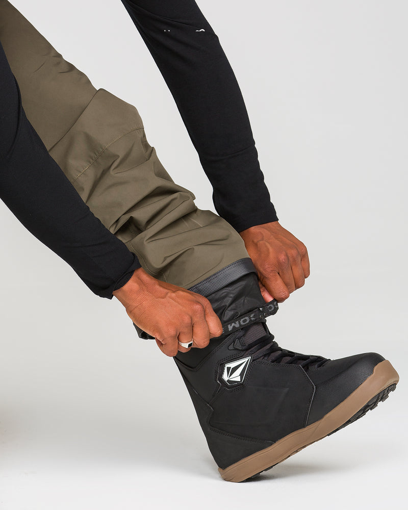 A person wearing Volcom Men’s Dua Gore-Tex Pants in olive-green adjusts the strap on a black snowboarding boot with a brown sole, ensuring waterproof protection and comfort. The background is plain white.