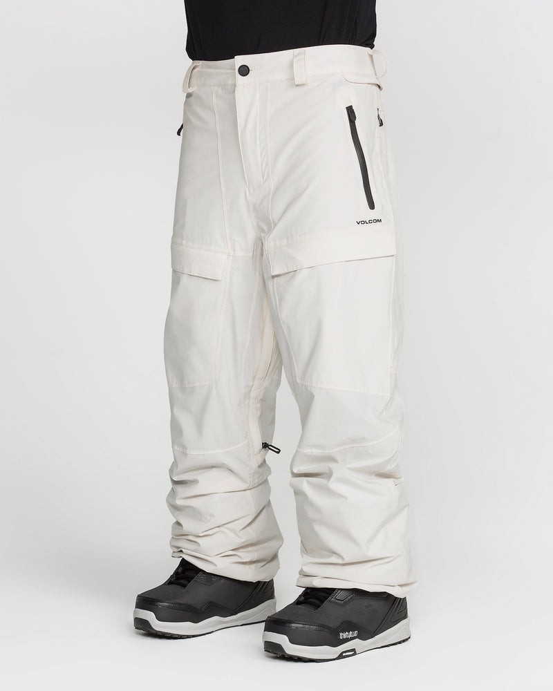 A person is wearing Volcom Men’s Rnge Stretch 30K Pants in off-white, featuring black zippers and buttons, along with Volcom branding on the right thigh. These baggy fit snow pants offer a functional 4-way stretch design and are paired with black and white winter boots, all set against a plain white background. The upper body is not visible.
