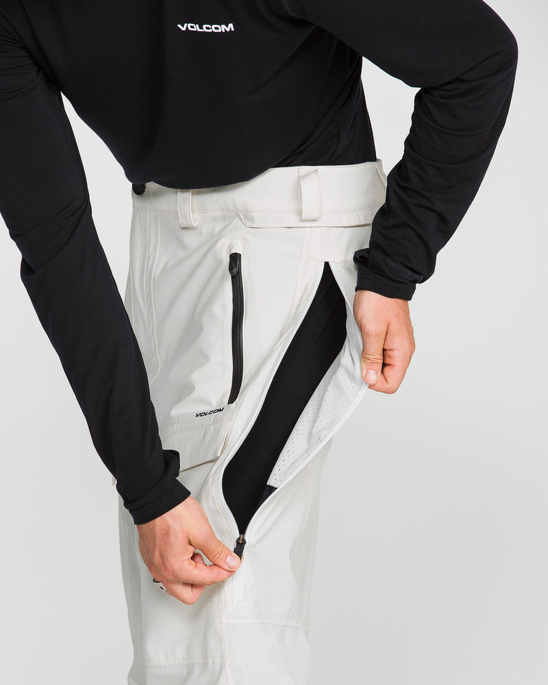 Wearing a black long-sleeve shirt with a VOLCOM logo on the chest, a person unzips the side vent of their Volcom Men’s Rnge Stretch 30K Pants in white, revealing the black inner lining. The baggy fit of the pants complements the shirt for a stylish look.