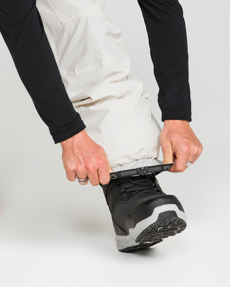 A person wearing Volcom Men’s Rnge Stretch 30K Pants with a baggy fit and black boots adjusts a strap at the bottom of their pant leg, securing it over the boot.