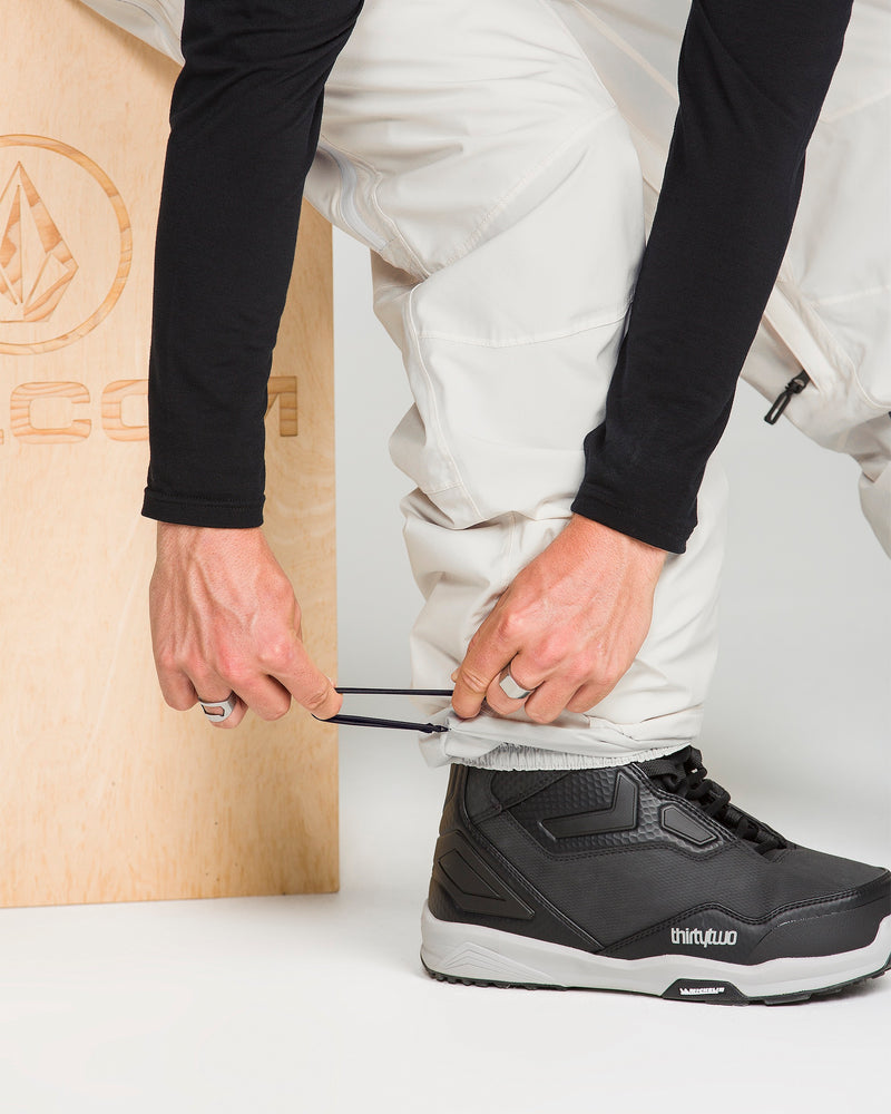 Wearing Volcom Men’s Rnge Stretch 30K Pants in white with a baggy fit, someone adjusts the ankle drawstring while sporting black ThirtyTwo snowboard boots beside a wooden box featuring the Volcom logo.