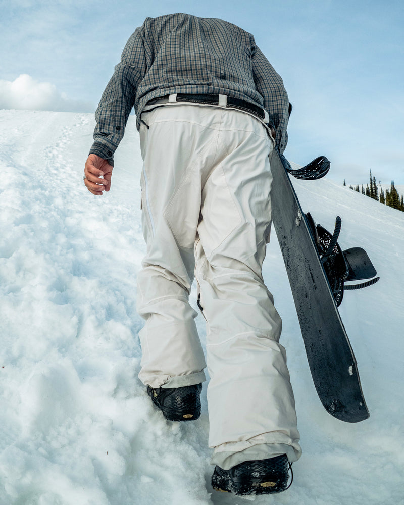 A person in Volcom Men’s Rnge Stretch 30K Pants and boots walks uphill on snow, carrying a snowboard in one hand with a clear sky in the background.