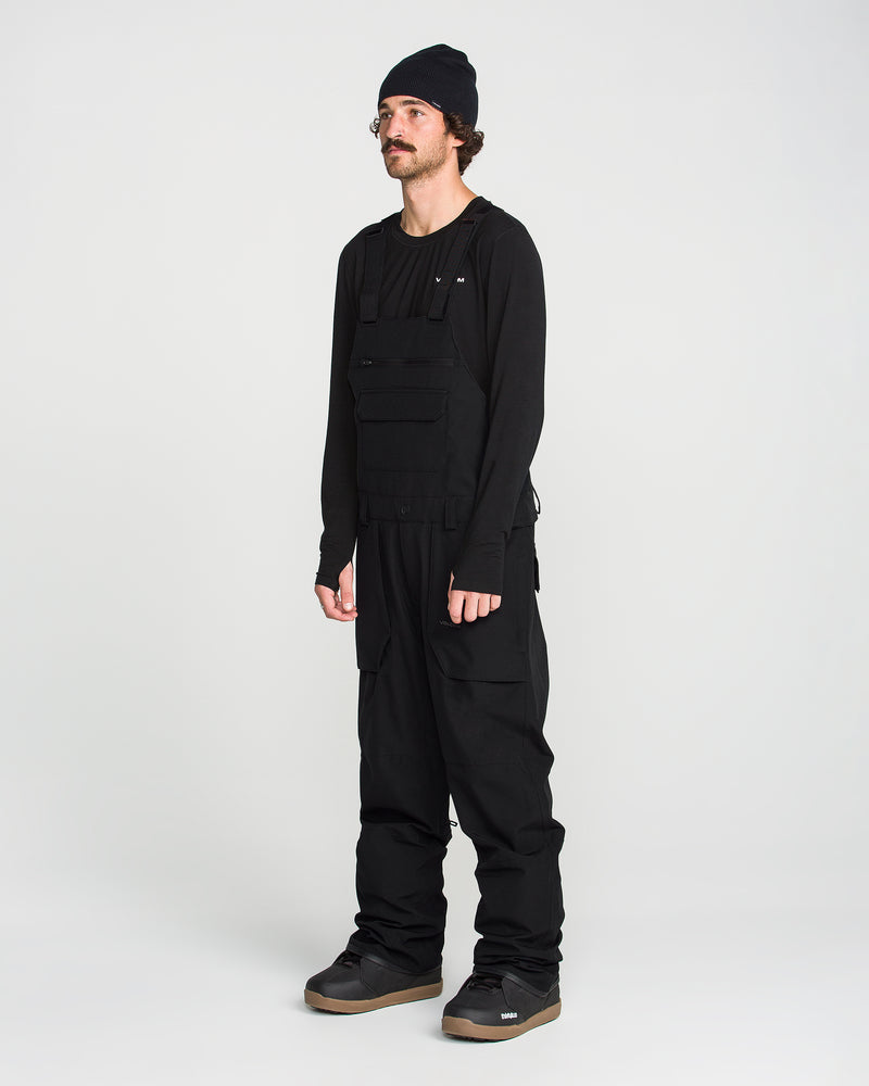 A person wearing a black beanie, long-sleeve shirt, utility vest, and the Volcom Men’s Roan Bib with adjustable suspenders stands against a plain white background, completing the look with gray sneakers.