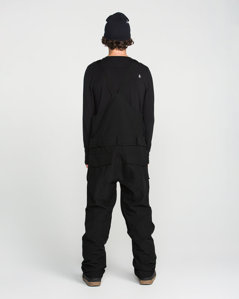 A person stands facing away, wearing a black beanie, a long-sleeve shirt, and the Volcom Men’s Roan Bib—a black recycled bib overall with adjustable suspenders—against a plain white background.