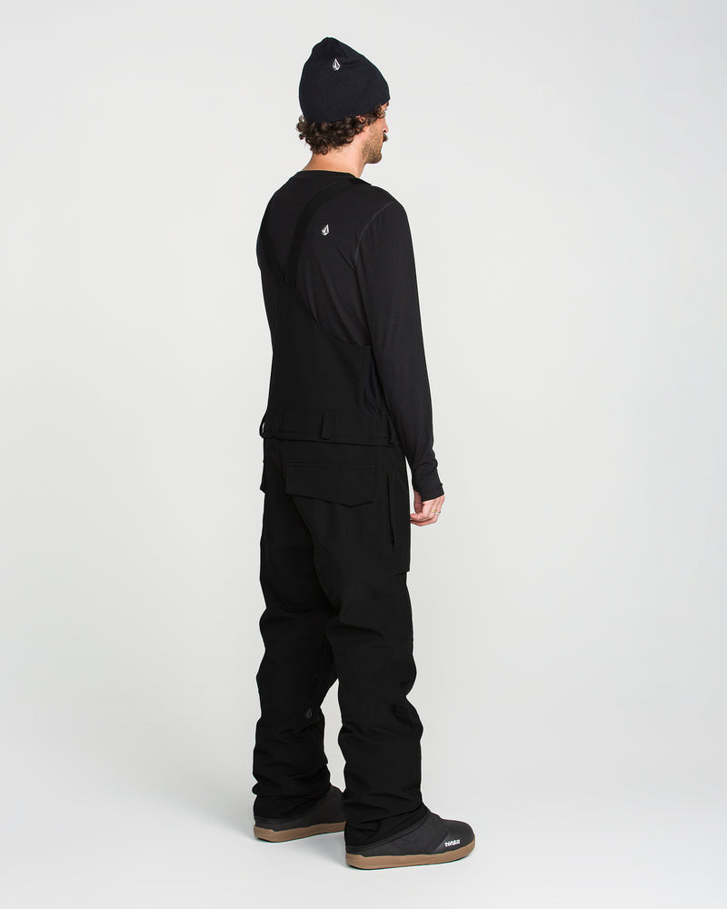 A person stands facing away, wearing a Volcom Men’s Roan Bib—a baggy recycled bib overall with adjustable suspenders—paired with a black beanie, long-sleeve shirt, and boots against a plain white background.