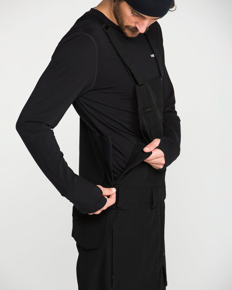 A person wearing a black long-sleeve shirt and the Men’s Roan Bib by Volcom, featuring adjustable suspenders, adjusts the side of their outfit against a plain white background.