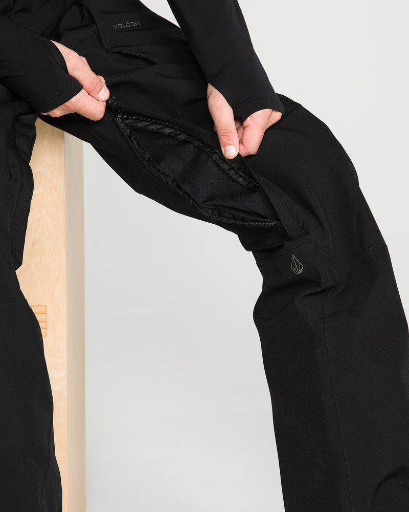 A person unzips the inner thigh vent of the black Volcom Men’s Roan Bib while seated on a wooden bench, highlighting its ventilation feature.