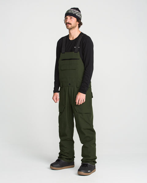 A man stands against a plain white background wearing a black long-sleeve shirt, Volcom Men’s Roan Bib crafted from recycled stretch Oxford for enhanced comfort and mobility, black shoes, and a patterned beanie. The dark green Volcom overalls feature 15K protection, delivering both style and functionality.