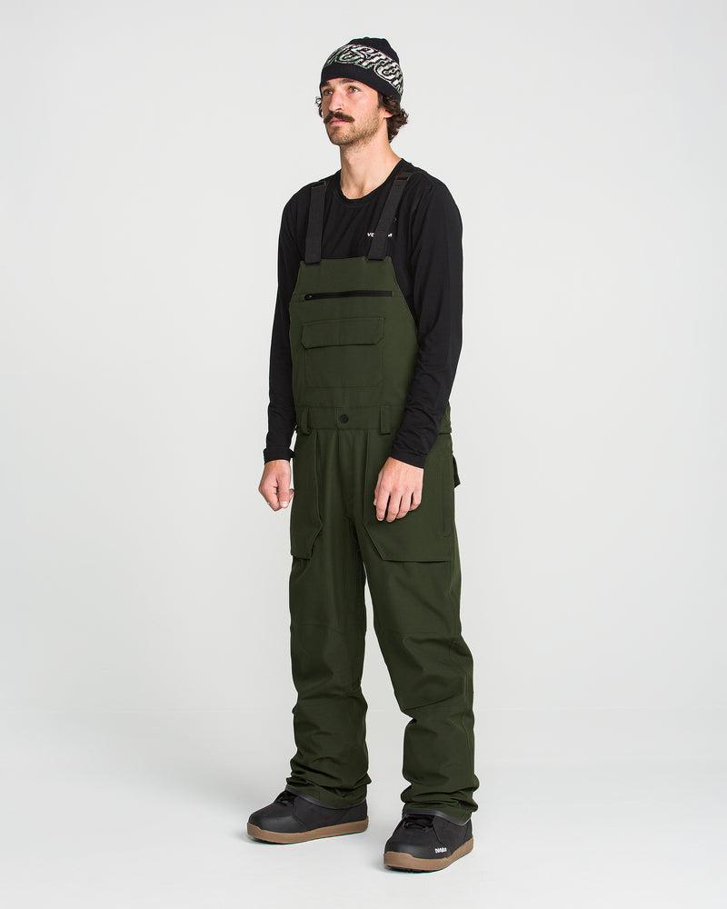 A man stands against a plain white background wearing a black long-sleeve shirt, Volcom Men’s Roan Bib crafted from recycled stretch Oxford for enhanced comfort and mobility, black shoes, and a patterned beanie. The dark green Volcom overalls feature 15K protection, delivering both style and functionality.