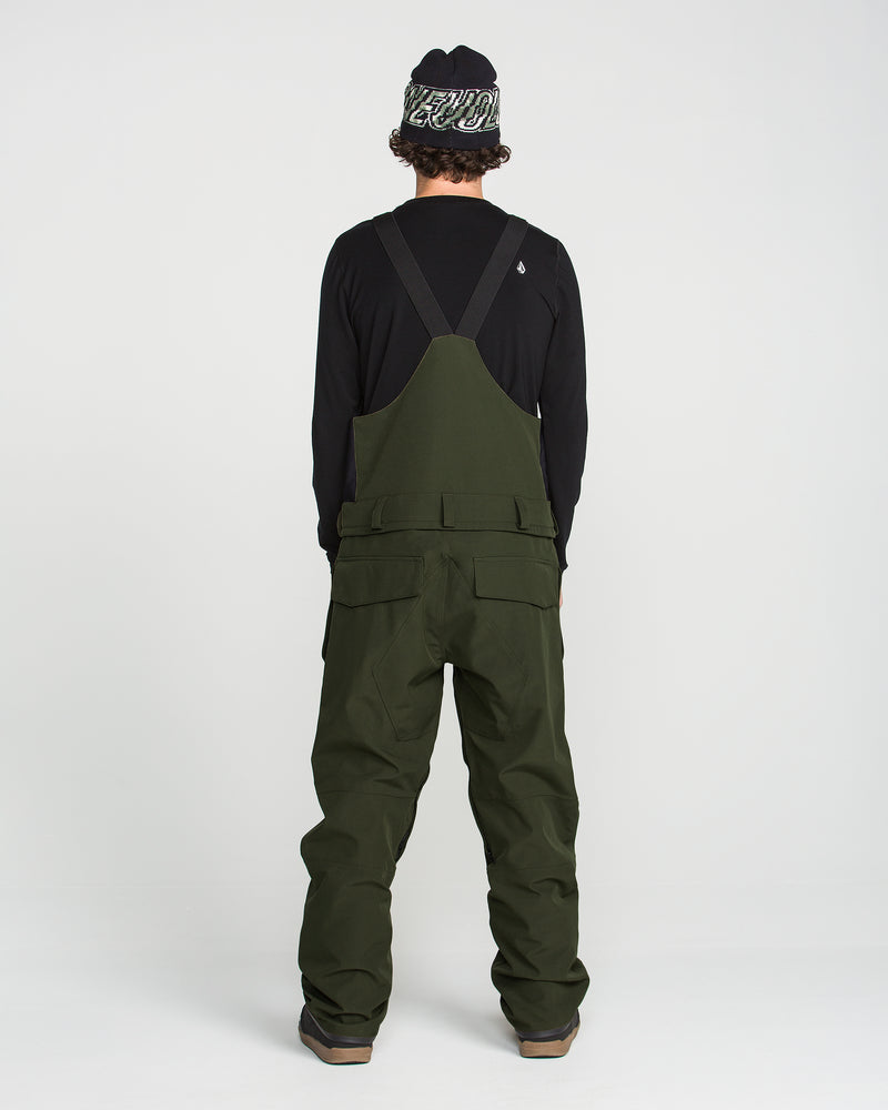 A person stands facing away, wearing the Volcom Men’s Roan Bib in dark green—crafted from recycled stretch Oxford fabric with 15K protection—paired with a black long-sleeve shirt and a black-and-white beanie. The plain white background highlights the overalls.