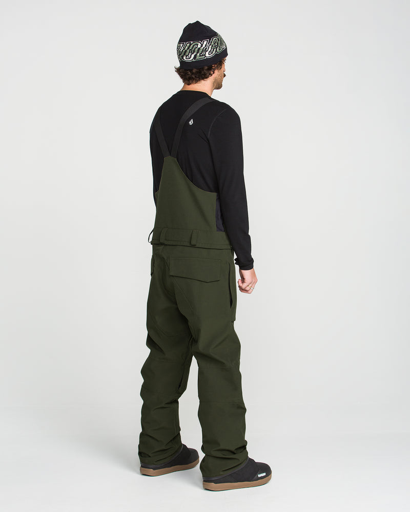 A person stands facing away, wearing a black long-sleeve shirt and the Volcom Men’s Roan Bib in olive green—expertly crafted from recycled stretch Oxford for comfort and durability—paired with black shoes and a black beanie with white writing, all set against a plain white background.