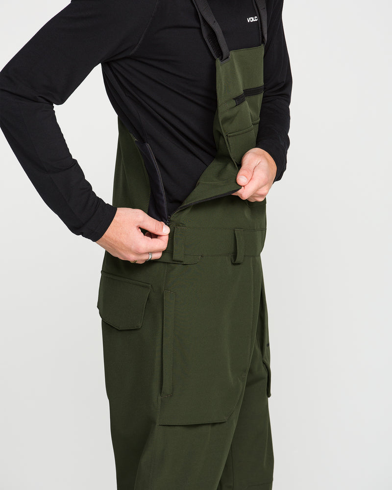A person is shown zipping up the side of Volcom Men’s Roan Bib Overalls in dark green, crafted from recycled stretch Oxford fabric with dependable 15K protection. The overalls are paired with a black long-sleeve shirt, and the individual’s head is not visible in the image.