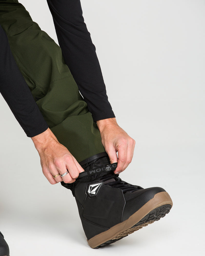 Wearing the Volcom Men’s Roan Bib in green and a black long-sleeve shirt, a person tightens the strap on a black snowboarding boot with a tan sole, ready for action with 15K protection and durable recycled stretch Oxford fabric.